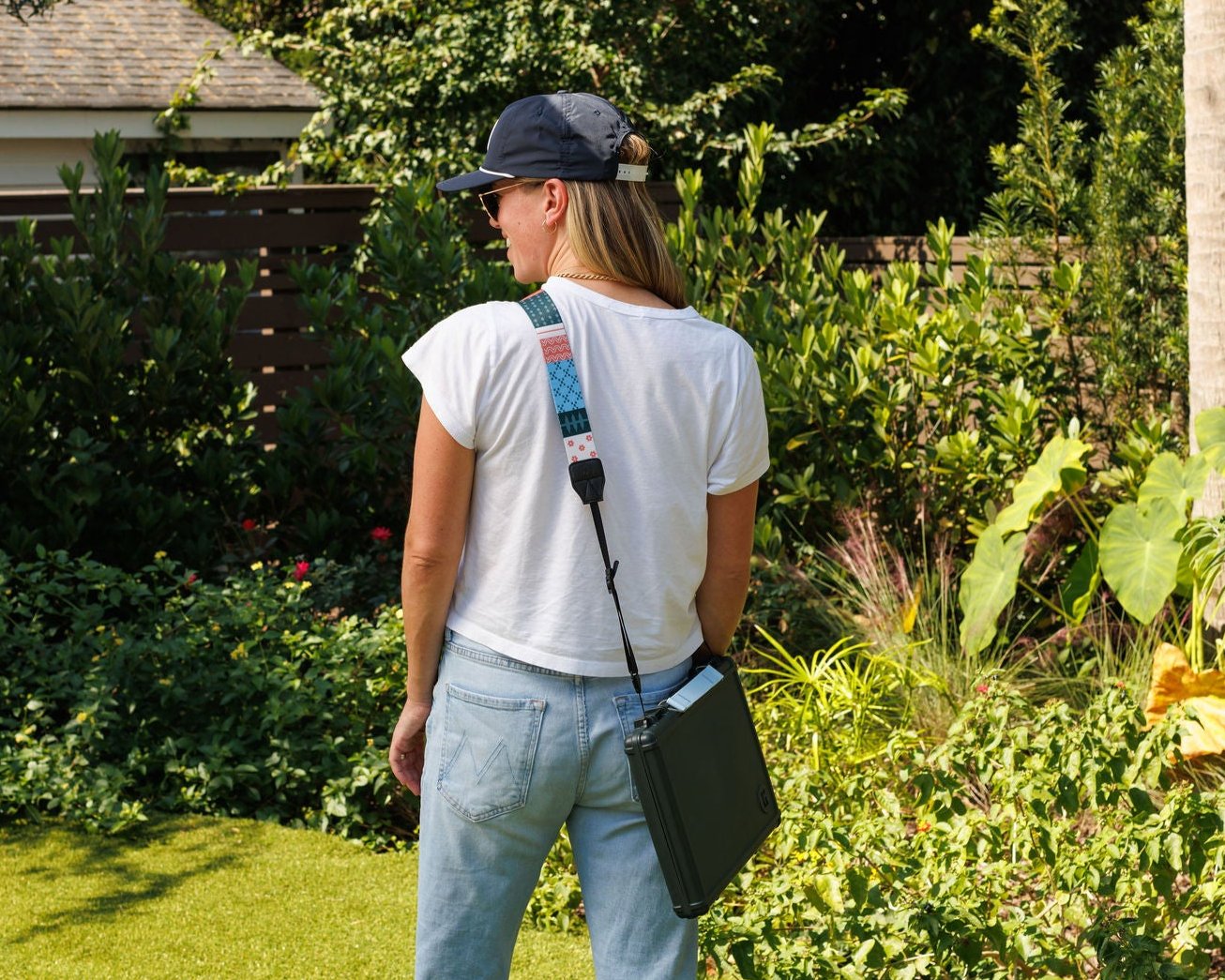 Woman in white shirt and hat carrying a portable backgammon board with strap in a garden