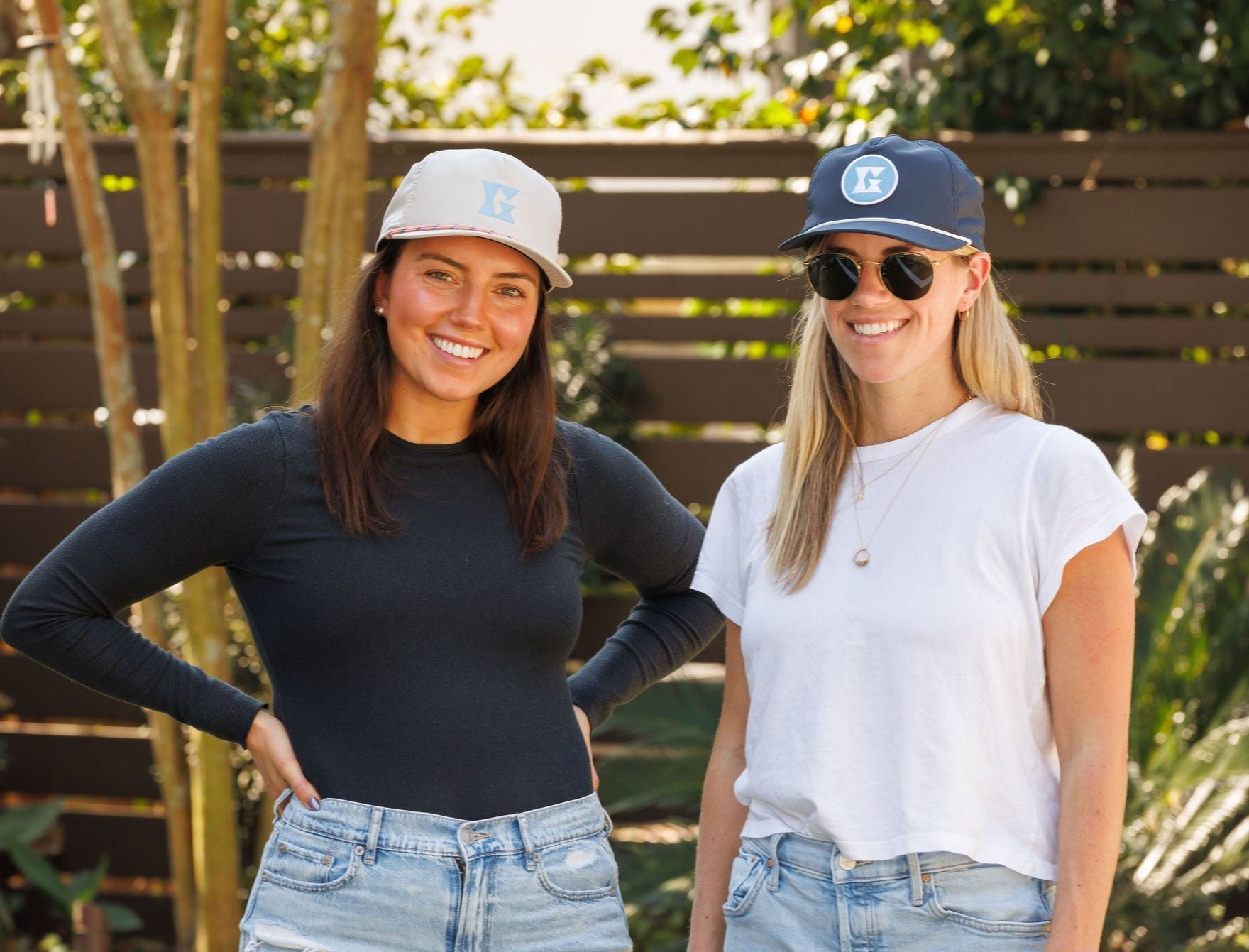 Two women smiling and wearing Gammn hats; one in black long-sleeve and the other in white t-shirt, standing outdoors with wooden fence and greenery