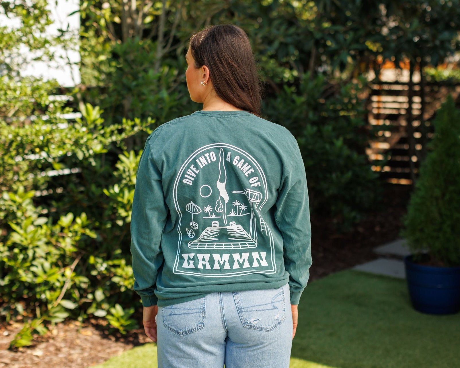 Woman wearing teal long-sleeve Gammn shirt with white “Dive into a game of” graphic and backgammon design on the back, standing outdoors among green plants and wooden fence.