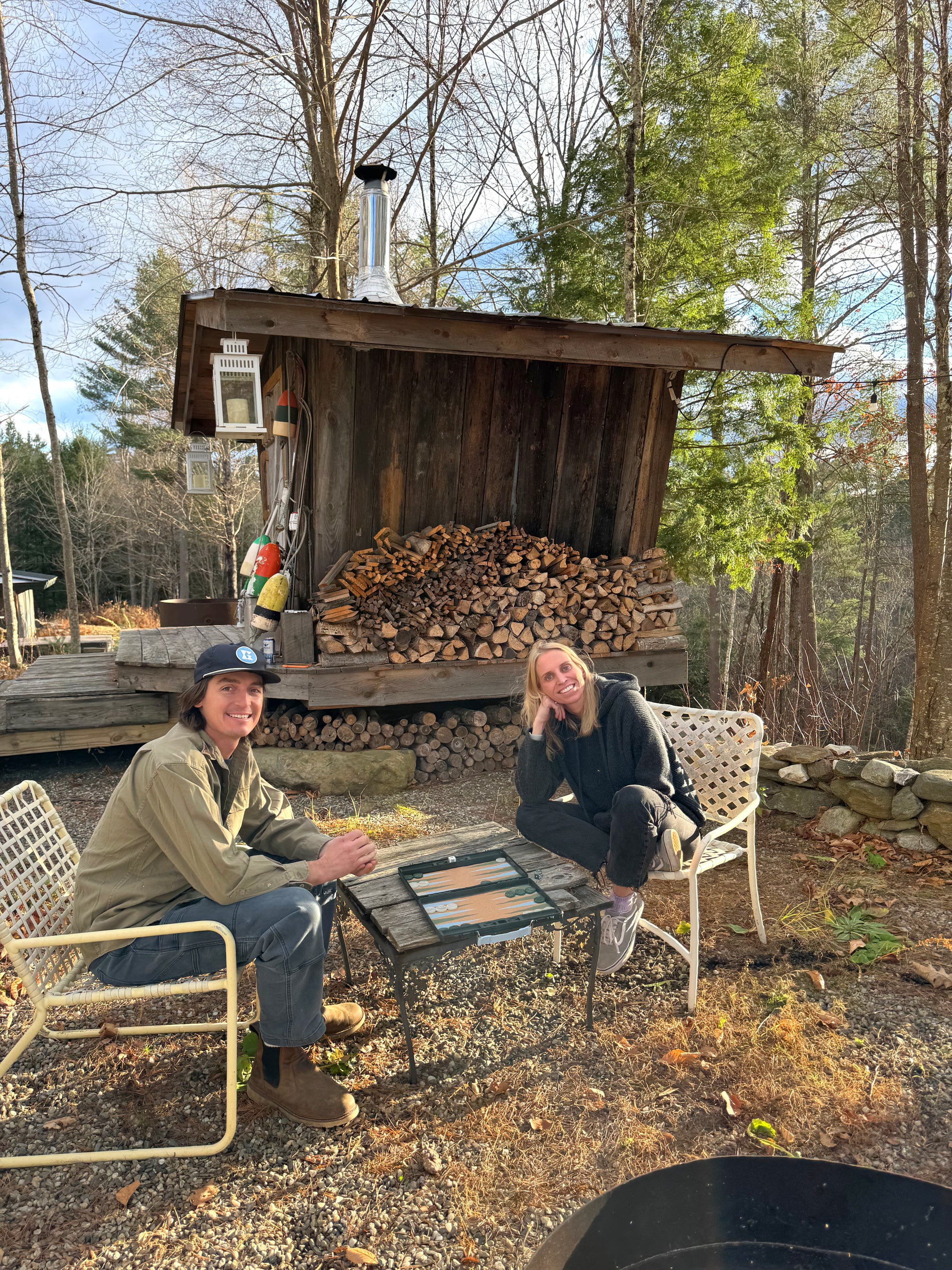 Two people playing a portable GAMMN backgammon set outdoors by a woodpile and forest – cozy cabin setting for travel board games
