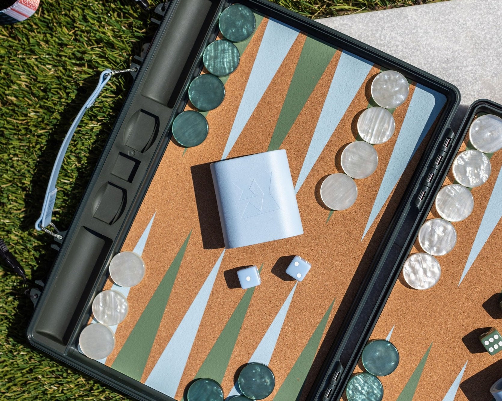 Open backgammon board on grass with orange cork playing surface and alternating green and white triangles, translucent green and white checkers, light blue dice and cup, and scoreboard